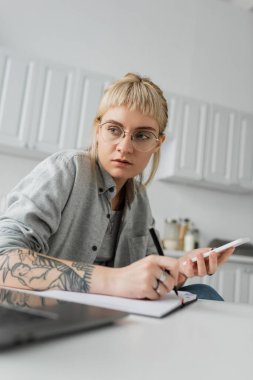 Eline dövme yaptırmış genç bir kadın deftere not alıyor, akıllı telefon ve kalemi beyaz masada tutuyor, ön planda bulanıklık var, evden çalışıyor. 