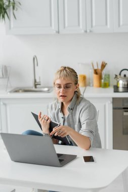 Elinde dövmesi ve elinde kâkülleri olan genç bir kadın. Akıllı telefonun yanında, ekranı boş. Beyaz masadaki laptopa bakıyor. 