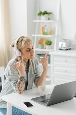 young woman with bangs and tattoo on hand sitting in wireless headphones and gesturing near laptop, cup of coffee and blurred smartphone with blank screen on table, freelance, work from home clipart