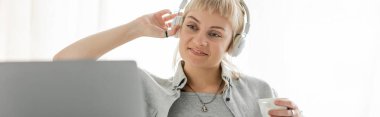 smiling young woman with bangs sitting in wireless headphones and holding cup of coffee while looking at blurred laptop at home, freelance, work from home, banner  clipart