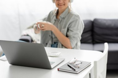 eyeglasses and pen on top of notebook near laptop and cheerful freelancer smiling while holding cup of coffee on blurred background at home, work from home, modern workspace  clipart