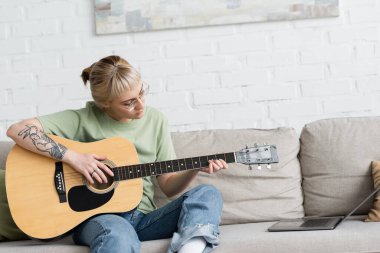 Kâküllü ve dövmeli genç bir kadın elinde akustik gitar tutarken ve bilgisayar başında video dersi alırken ve evdeki modern oturma odasında rahat bir koltukta otururken çalmayı öğrenirken.