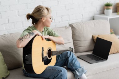 Kaküllü, dövmeli, akustik gitarlı genç bir kadın. Dizüstü bilgisayarında boş ekran ve oturma odasında rahat bir kanepede otururken gitar çalmayı öğreniyor.