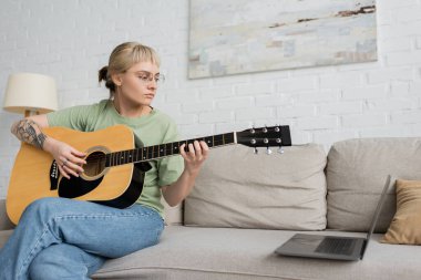 Kaküllü ve dövmeli genç bir kadın akustik gitar çalarken dizüstü bilgisayar başında video dersi alıyor ve evdeki modern oturma odasında rahat bir koltukta oturuyor. 