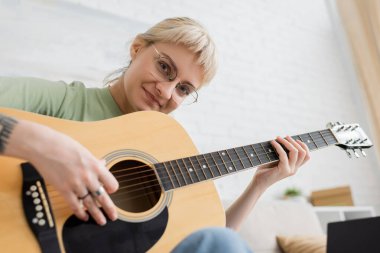 Elinde kâkülleri ve dövmesi olan neşeli genç bir kadın akustik gitar çalıyor ve modern oturma odasında oturmuş müzik öğrenirken kameraya bakıyor.