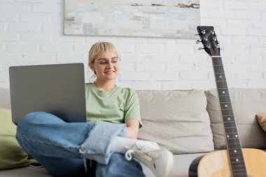 Sarışın, kısa saçlı, kâküllü ve gözlüklü mutlu genç bir kadın rahat bir koltukta oturup duvarında resim olan gitara bakarken dizüstü bilgisayar kullanıyor. 