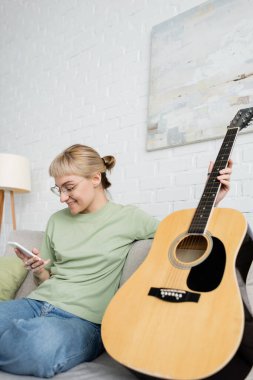 Sarışın, kısa saçlı, perçinli, gözlüklü, akıllı telefon kullanan mutlu genç bir kadın modern oturma odasındaki gitarın yanında, duvarında resim olan rahat bir kanepede oturuyor. 