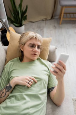 Dövmeli genç bir kadın, sarışın ve kısa saçlı, perçem ve gözlüklü, akıllı telefon kullanan modern oturma odasındaki rahat koltukta dinlenen, bulanık arkaplan.