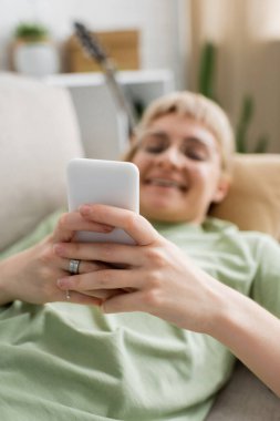 Dövmeli mutlu bir genç kadın, sarışın ve kısa saçlı, perçem ve gözlüklü akıllı telefon kullanan modern oturma odasında rahat bir kanepede dinlenen bulanık bir arka plan.