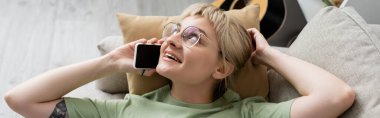 Sarışın, kısa saçlı, perçinli, gözlüklü, akıllı telefondan konuşan modern oturma odasındaki rahat koltukta dinlenen mutlu genç kadın manzarası. 