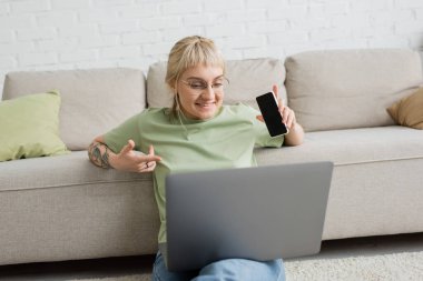 cheerful and tattooed woman with bangs and eyeglasses using laptop while sitting on carpet and holding smartphone with blank screen near comfortable couch in modern living room  clipart
