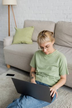 tattooed woman with bangs and eyeglasses using laptop while sitting on carpet near smartphone with blank screen and comfortable couch in modern living room  clipart