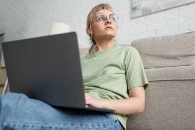 Düşük açılı sarışın, kısa saçlı, kâküllü ve gözlüklü bir kadının modern oturma odasında rahat bir kanepenin yanında otururken ve duvarda para öderken dizüstü bilgisayar kullanması. 