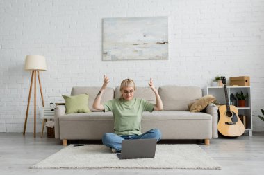 emotional tattooed woman with bangs and eyeglasses using laptop while sitting on carpet near smartphone, comfortable couch, guitar and rack with plants in modern living room with paiting on wall  clipart