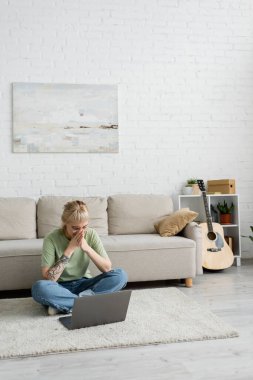 Kaküllü, gözlüklü, ağzını kapatan, dizüstü bilgisayarı olan, gitar ve rafların yanındaki rahat koltukta oturan, modern oturma odasındaki bitkilerle duvara asılan, dövmeli kadın. 