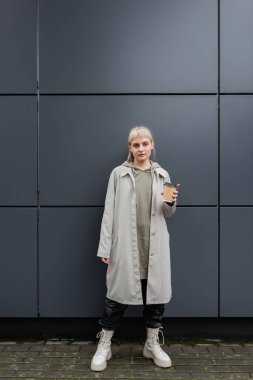 stylish young woman with blonde hair with bangs standing in coat and hoodie while holding paper cup with coffee and looking at camera near grey modern building on street, outside, urban living  clipart