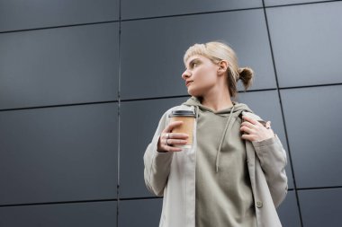 Havalı, sarı saçlı, kâküllü, ceketli ve kapüşonlu bir kadın. Elinde kahve bardağıyla caddede, dışarıda, kentte yaşayan gri modern binanın yanında duruyor. 