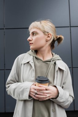 stylish young woman with blonde hair with bangs standing in coat and hoodie while holding paper cup with takeaway coffee near grey modern building on street, outside, urban living, look away  clipart