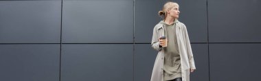 stylish young woman with bangs standing in coat and hoodie while holding paper cup with takeaway coffee near grey modern building on street, outside, urban living, look away, banner clipart