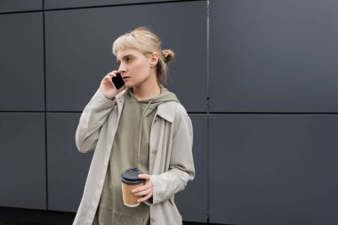 Kâküllü, sarı saçlı, elinde kahve bardağıyla şık bir genç kadın. Akıllı telefonla konuşurken, kapüşonlu ve ceketli bir kadın. Şehir sokağındaki gri modern binanın yanında duruyor. 