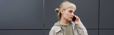 stylish young woman with bangs and blonde hair talking on smartphone and standing in hoodie and coat near grey modern building on urban street, banner, looking away, urban lifestyle  clipart