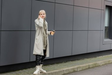 fashionable young woman with bangs holding paper cup with coffee to go while adjusting wireless earphones and standing in trendy outfit, hoodie and coat near grey modern building on street outside  clipart