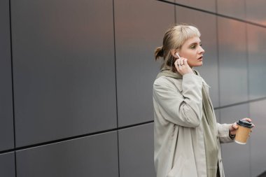 blonde woman with bangs holding paper cup with coffee to go while adjusting wireless earphones and standing in trendy outfit, near grey modern building on urban street, look away  clipart