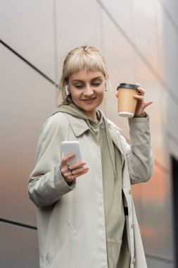 happy young woman with bangs holding paper cup with coffee to go and standing in trendy outfit and wireless earphones while using smartphone near grey modern building on urban street  clipart