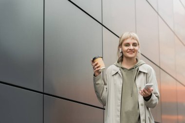 smiling young woman with bangs holding paper cup with coffee to go and standing in trendy outfit and wireless earphones while using smartphone near grey modern building on urban street, look at camera clipart