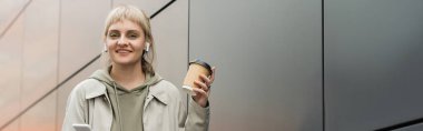 Elinde kahküllü, elinde kahve bardağıyla gülümseyen genç bir kadın modaya uygun kıyafetler ve kablosuz kulaklıklarla şehir sokağındaki gri, modern binanın yanında akıllı telefon kullanıyor.