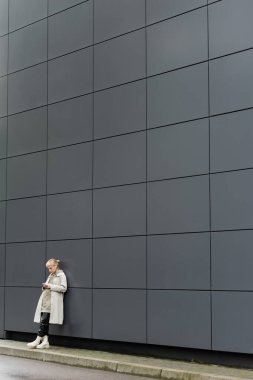 Paçalı elbiseleri ve kablosuz kulaklıkları olan genç bir kadın şehir sokağındaki gri modern binanın yanında akıllı telefon kullanırken kapüşonlu, siyah deri pantolonlu ve botlu bir kadın.
