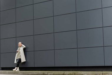 Kâküllü genç bir kadın şık kıyafetleri ve kablosuz kulaklıkları ile şehir sokağındaki gri modern binanın yanında akıllı telefon kullanırken kapüşonlu, siyah deri pantolonlu ve botlu.