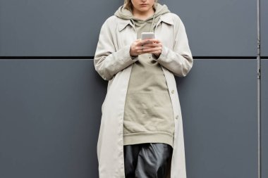 Modayı takip eden genç bir kadının moda kıyafetleri içinde durduğu ve şehir sokağındaki modern binanın gri duvarının yanında akıllı telefonuyla mesajlaştığı kapüşonlu ceket, kısmi görüş.