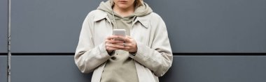 Modayı takip eden genç bir kadının moda kıyafetleri içinde durduğu ve şehir sokağındaki modern binanın gri duvarının yanında akıllı telefonla mesajlaştığı kapüşonlu ceket, kısmi manzara, afiş. 
