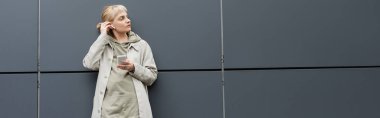 Havalı kıyafetleri ve kablosuz kulaklıkları olan şık, genç bir kadın. Şehir sokağındaki gri modern binanın yanında akıllı telefon kullanıyor. Kapüşonlu bir ceket, başka tarafa bakıyor. 