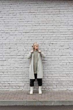 young woman with makeup, blonde hair, bangs, in stylish outfit, long hoodie, black leather pants and beige boots adjusting coat and standing near grey brick wall of modern building while looking away  clipart