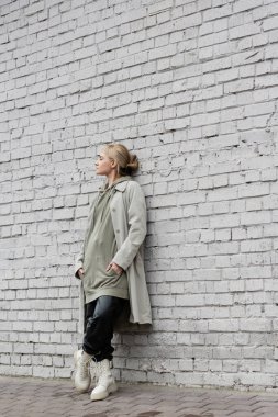 young woman with makeup, blonde hair, bangs, in stylish outfit with long hoodie, coat, black leather pants and beige boots standing with closed eyes near grey brick wall of modern building  clipart