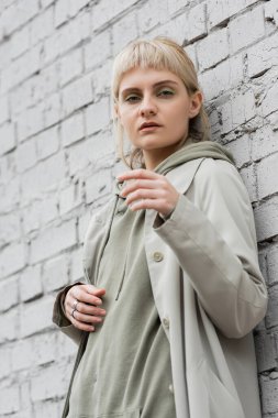 young woman with makeup, blonde hair, bangs, in stylish outfit standing near grey brick wall of modern building and looking at camera while posing outdoor, coat, fashion trend  clipart