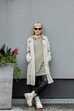 full length of young woman in stylish sunglasses, long hoodie, coat, black leather pants and beige boots standing near grey wall of modern building and posing with hands in pockets near green plant  clipart