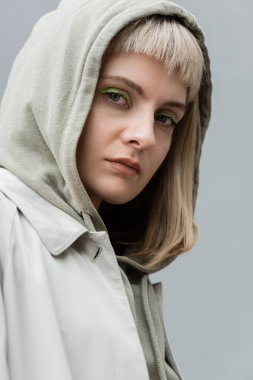 stylish and young woman with bangs and blonde hair standing with hood on head and trendy comfortable clothes while looking at camera isolated on grey background in studio, hoodie  clipart