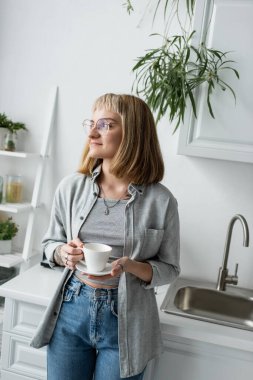 Kısa saçlı, kâküllü, gözlüklü, dövmeli, elinde kahve fincanı olan mutlu genç bir kadın mutfak dolaplarının ve modern apartmandaki bitkilerin yanında günlük gri giysiler içinde dikiliyor. 