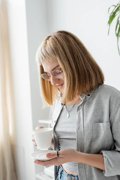 stock image happy young woman with short hair and bangs, eyeglasses necklace, ring and tattoo holding cup of morning coffee and saucer while standing in casual grey clothes and smiling in modern kitchen 