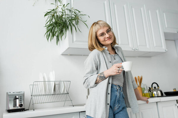 happy young woman with short hair and bangs, eyeglasses and tattoo holding cup of morning coffee while standing in casual clothes next to toaster, dishes, kettle and plant in modern kitchen 