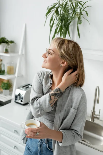 happy young woman with bangs, eyeglasses and tattoo on hand adjusting short hair and holding cup of morning coffee while standing in casual clothes next to toaster, kitchen sink and faucet at home