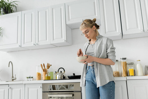 young woman with bangs, eyeglasses and short hair holding bowl with breakfast and spoon while standing in casual grey clothes next to kettle, kitchen appliances and blurred white cabinets at home 
