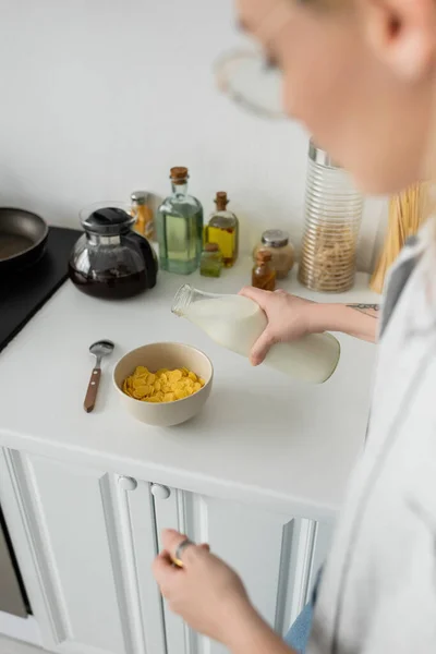 blurred and tattooed young woman in eyeglasses holding bottle while pouring fresh milk into bowl with cornflakes on kitchen worktop while making breakfast and standing in modern kitchen