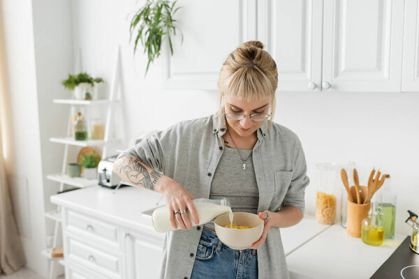 tattooed young woman with bangs in eyeglasses holding bottle while pouring fresh milk into bowl with cornflakes and making breakfast while standing in casual clothes in modern kitchen 