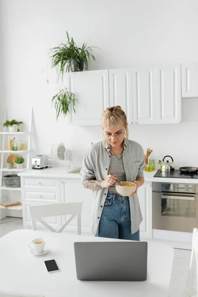 tattooed young woman with bangs, eyeglasses and short hair holding bowl with cornflakes while having breakfast and looking at laptop near smartphone and cup of coffee on desk in modern kitchen