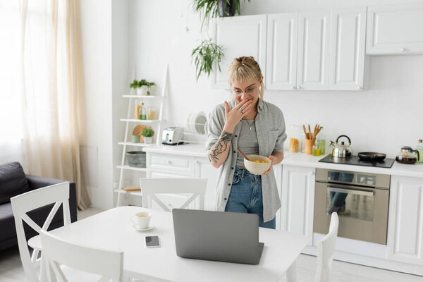 татуированная молодая фрилансер с челкой, очки и короткие волосы покрывающий рот во время смеха, держа миску с кукурузными хлопьями и глядя на ноутбук возле смартфона и чашку кофе на столе на кухне 