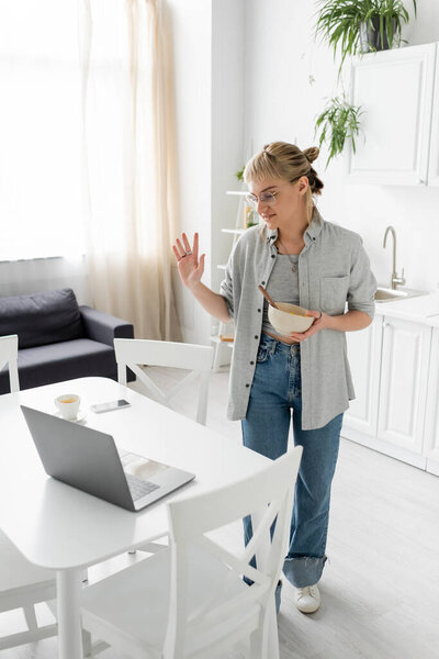 татуированный молодой фрилансер с челкой и очками, держащий миску с кукурузными хлопьями, глядя на ноутбук и размахивая рукой во время видеозвонка возле смартфона и чашки кофе на столе на современной кухне 